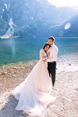 Beautiful wedding couple tenderly hug on the background of mountains and the river, long white dress of the bride. clipart