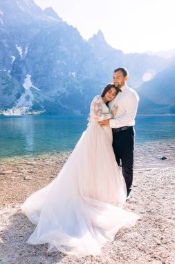 Romantik bir an. Gülümseyen yeni evliler, gelin ve damat dağlarda güzel bir göl kenarında kucaklaşıyorlar.