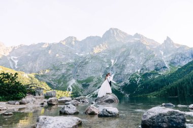 Gelin, beyaz gelinlik içinde damada arkadan sarılıyor, göl kenarındaki bir taşın üstünde, dağ manzaralı.
