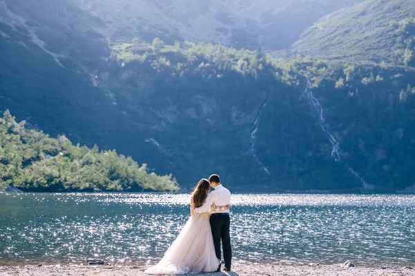 新婚夫妇背对着站在那里 看着美丽的湖水 新娘和新郎拥抱在沙滩上 — 图库照片