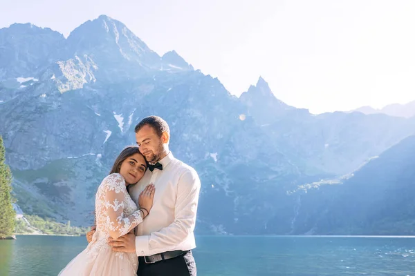 美しい白いドレスを着た花嫁と美しい緑の山々と青い水の湖を見下ろす新郎 — ストック写真