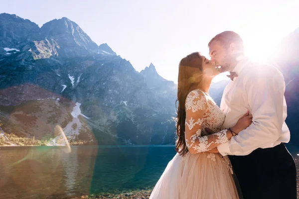 快乐而年轻的新婚夫妇在湖边亲吻 有一个新郎和一个长卷发的漂亮的新娘 在群山和光芒的背景下 — 图库照片