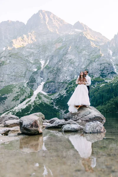 新娘和新郎在山中湖边 在山水的背景下 新婚夫妇在一起 浪漫的一对恋爱中的夫妻站在石头海岸上 风景秀丽的山景 — 图库照片
