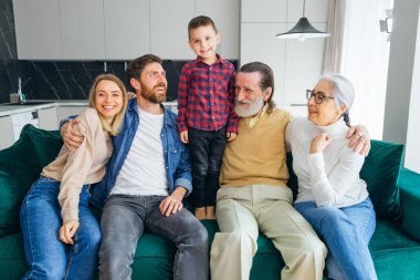 Sıcak oturma odasında kameraya gülümseyen mutlu bir aile. Neşeli bir adam ve kadın, çocuklar ve ebeveynlerle birlikte kanepede oturuyorlar. Kendi Nesillerinde Büyükbabalar, Ebeveynler ve Çocuklar
