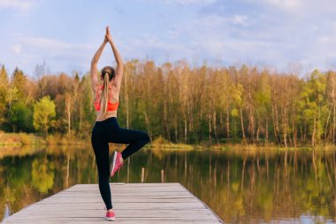 Vrikshasana pozu giymiş arka görüşlü kadın, dışarıda yoga yapıyor. Refah, sağlık konsepti. Göl İskelesi