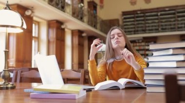 Yorgun ve bitkin bir öğrenci kütüphanede kitap ve dizüstü bilgisayarla oturuyor, sınava hazırlanıyor, ara veriyor, gözlüklerini çıkarıyor ve boynunu geriyor..