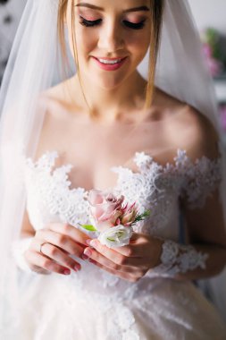 Bride in a beautiful dress standing indoors, home interior. Fashionable wedding style. Wedding makeup and hairstyle. Beautiful woman holding a boutonniere