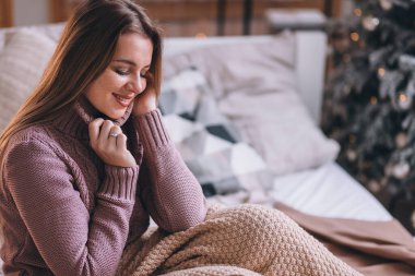 Sıcak bir süveter giyen güzel bir kız rahat bir odada Noel ağacının arka planında oturuyor. Yeni Yıl