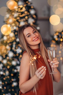 Modaya uygun bir iç mekan, bluzlu güzel bir kadın, süslü bir Noel ağacının yanında poz veriyor, elinde şampanya ve maytap var..