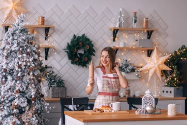Genç bir kadın Noel için kurabiye yapıyor. Oturma odası arka planda Noel ağacı süslemeleri ile süslenmiş. Önlüklü kadın..