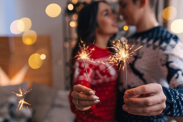 Пара влюбленных в теплой одежде в свитерах красного и белого цвета обнимаются и целуются, держа искры на фоне рождественской елки в украшенной студии
