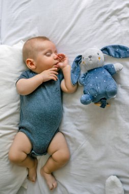 Bir bebek, parlak bir yatak odasında mavi bir takım elbiseyle yatakta baş parmağını emer, peluş tavşanla uzanan sevimli bir bebek.