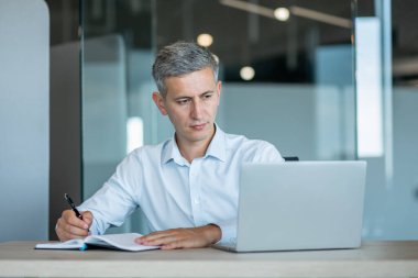 Gri saçlı profesyonel bir adam modern bir ofiste masa başında oturup dizüstü bilgisayarına konsantre olarak not alır. Şirket ortamında odaklanma, analiz ve iş verimliliği.