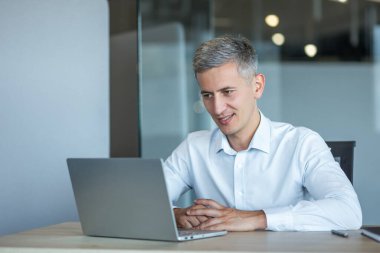 Kendine güveni tam, gri saçlı bir adam modern bir ofiste dizüstü bilgisayarıyla çalışıyor.