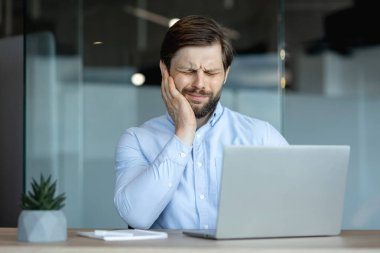 Mavi gömlekli bir işadamı, dizüstü bilgisayarı üzerinde çalışırken sinirli görünüyor. Diş ağrısını hisset