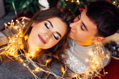 Üst kattaki kız gözlerini kapadı ve erkek ona sarıldı. Noel ağacının yakınındaki çelenklere sarılmış çiftler..