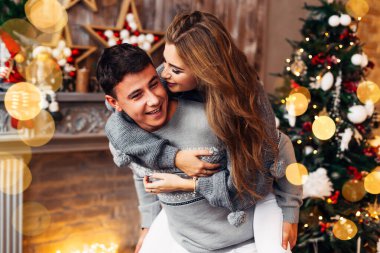 Kız arkadan adama sarılıyor ve onu öpmek istiyor. Birbirlerine aşık çiftler şöminenin ve Noel ağacının yanında.