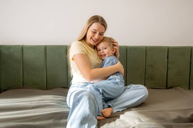 Açık sarı tişörtlü gülümseyen bir anne, kot elbiseli küçük çocuğuna sarılıyor. Yatakta sakin, sevgi dolu bir aile anı yaşıyor..