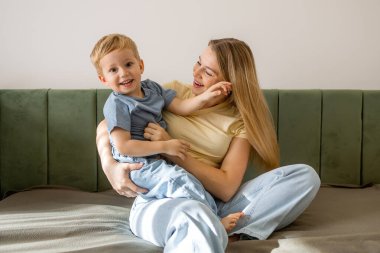 Sevgi dolu bir anne, gülümseyen oğlunu rahat bir yatakta sıcak, bağlanan ve günlük aile yaşamında rahat bir yuva ortamında tutar..