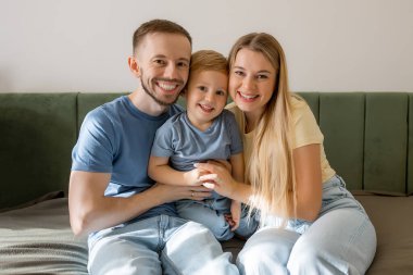 Neşeli bir aile yatakta birlikte poz verir, sıcak bir gülümseme paylaşır. Sıradan kıyafetler ve yumuşak doğal ışık.