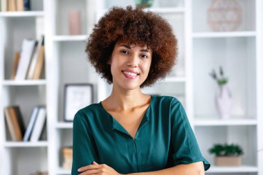 Afro saçlı, kendinden emin gülümseyen Afro-Amerikan kadın kollarını kavuşturmuş kameraya bakarak dostça gülümsüyor.