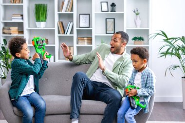 tapılası Afrikalı Amerikalı çocuklar plastik oyuncak tabancalarla oynuyorlar, ilgili genç babalarıyla, hafta sonları evde eğleniyorlar, kanepede oturuyorlar..