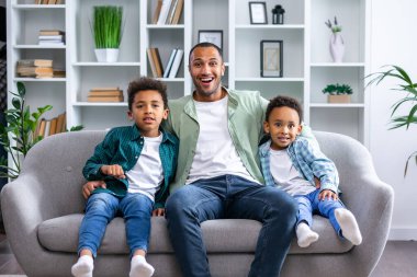 Küçük çocuklu mutlu bir aile oturma odasında kanepede oturur ve kameraya bakarak eğlenir. Gülümseyen baba kanepede dinleniyor. Küçük çocuklarla birlikte hafta sonunun keyfini çıkarın.