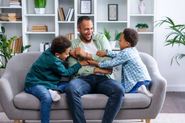 Babaları kanepede dinlenen çocuklar, küçük çocuklar eğleniyor, aile oturma odasında vakit geçiriyor.