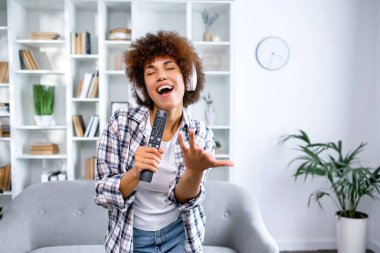 Mikrofon olarak uzaktan kumandalı kablosuz kulaklıklı güzel Afrikalı Amerikalı kadın Coronavirus karantinası sırasında evde en sevdiği şarkıları söylüyor.