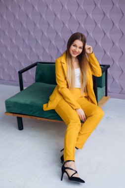 Uzun saçlı, sarı pantolonlu ve siyah topuklu ayakkabılı şık bir kız. Koltukta oturuyor..