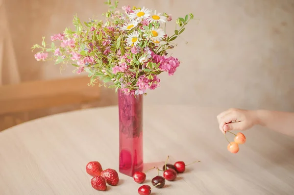 A bouquet of different rural flowers with camomiles in a pink vase on a table with ripe cherries. Summertime background.