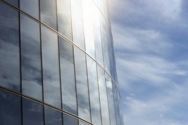 Mavi gökyüzü, güneş ve beyaz bulutlar modern bir iş merkezinin camlarına yansıyor.