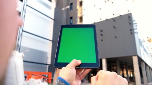 Tableta con pantalla verde sobre el fondo de un edificio en construcción. — Vídeos de Stock