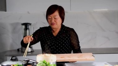 Asyalı kadın bambu buharı açıyor ve hamur köftelerini yemek çubuklarıyla tahta kesme tahtasına taşıyor.