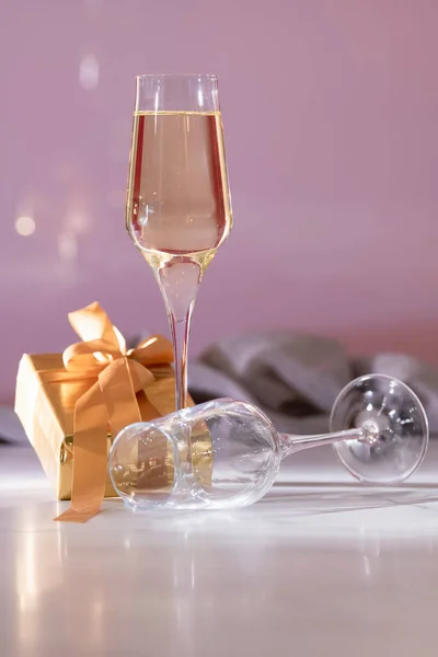 Champagne glass surrounded by a lying down empty glass and a golden wrapped gift box on an out of focus background. Celebration and gifts concept.