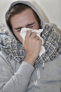 Fularlı ve kapüşonlu genç bir adamın kağıt peçeteyle burnunu sildiği yakın plan. Hastalık ve sağlık hizmeti kavramı.
