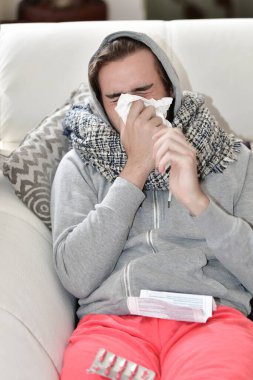 Kapüşonlu ve eşarplı genç bir adamın odak noktası dışında kağıt peçete kullanarak burnunu silmesi. Hastalık ve tıp konsepti.