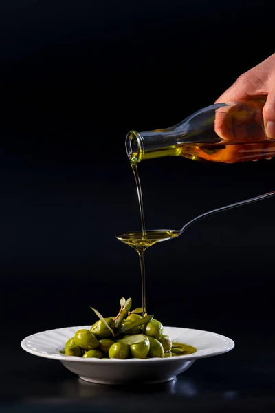 view of pure olive oil pouring on a metal spoon and overflowing into a plate with tempting olives