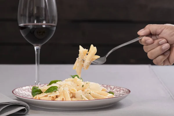 Enfoque selectivo de pasta recién cocida con queso en un tenedor sostenido por una mano sobre un plato con una copa de vino fuera de foco. Concepto de estilo de vida. — Foto de Stock