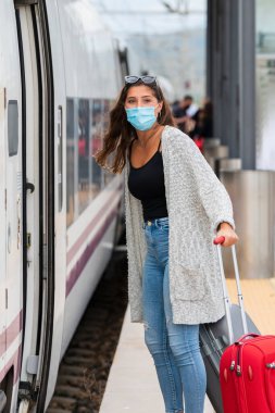 Genç bir kadın, kameraya bakarken trenin yanında iki bavul tutan cerrahi maske kullanıyor. Turizm ve güvenlik kavramı.