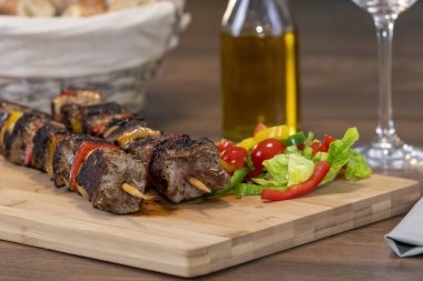 Tahta bir kesme tahtasının üzerinde çeşitli kesilmiş sebzelerin yanında iki biftek şişi.