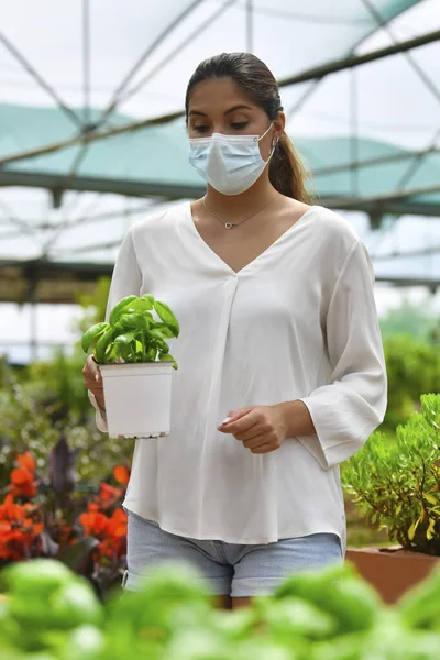 Mujer usando una máscara quirúrgica sosteniendo una maceta con una planta en un centro de jardinería — Foto de Stock