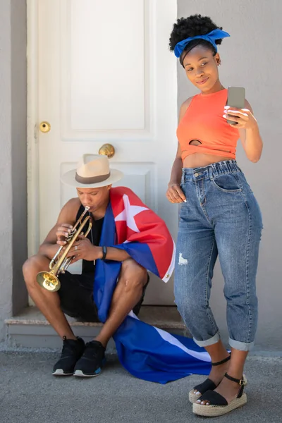 Mujer latina tomando una foto de sí misma junto a un hombre latino con una bandera cubana tocando una trompeta — Foto de Stock