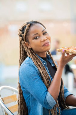 Afro-Amerikalı kadın elinde pizza dilimiyle kameraya gülümsüyor.