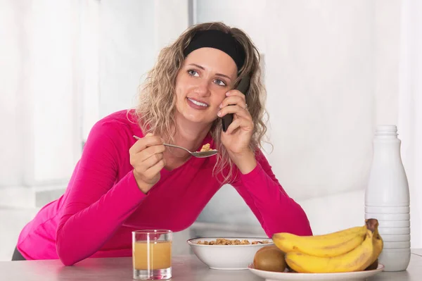 Mulher apoiada na mesa tomando café da manhã cereal com frutas e leite enquanto fala ao telefone — Fotografia de Stock