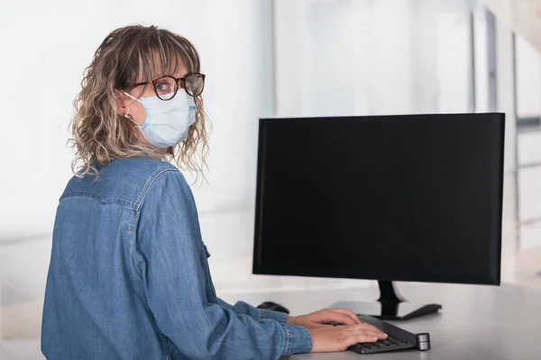 Vista trasera de una mujer adulta que usa mascarilla protectora en la oficina usando computadora con pantalla oscura para copiar espacio — Foto de Stock