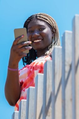 Telefonu kullanan ve önünde ahşap bir çit olan kameraya bakan, arkasında mavi gökyüzü olan kadın.