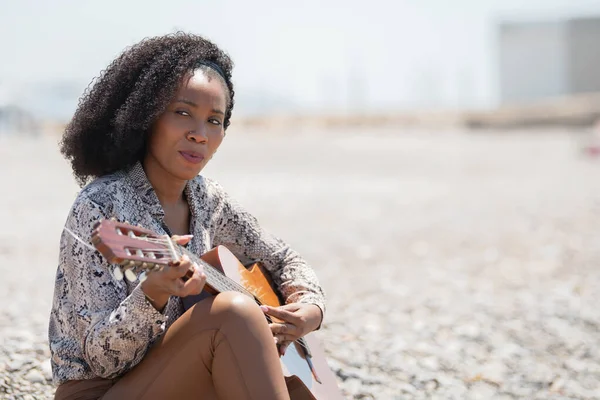 Młoda kobieta siedzi na żwirowej kamiennej ziemi na zewnątrz grając na gitarze akustycznej odwracając wzrok — Zdjęcie stockowe