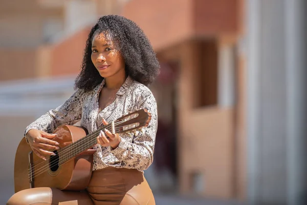 Mujer de pie tocando la guitarra mirando a la cámara sobre un fondo urbano desenfocado — Foto de Stock