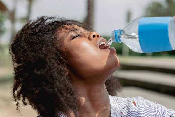 Afro-Frau trinkt Wasser aus einer Flasche, während sie an einem heißen und sonnigen Tag im Freien spaziert. lizenzfreie Stockbilder